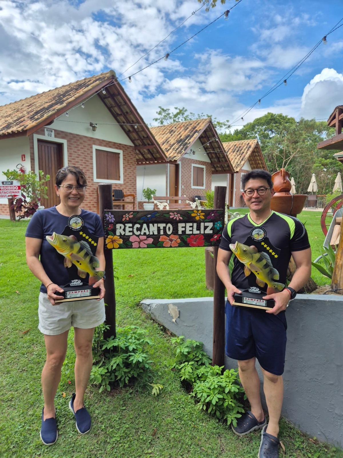 Casal com troféus em frente à placa do Recanto Feliz